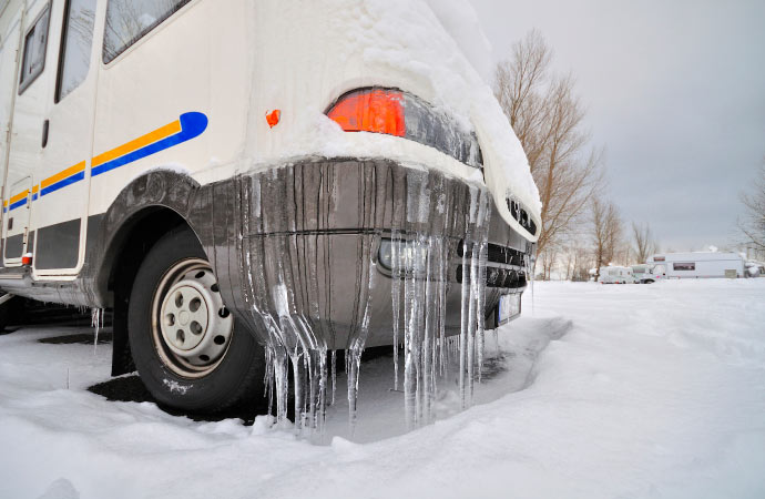 Icicles hang from the bumper and wheel well of a snow-covered white RV van.